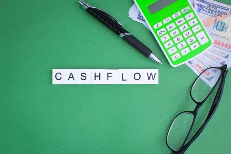 Scrabble-style letter tiles spelling CASHFLOW beside a calculator, hundred-dollar bills, eyeglasses, and a pen on a green background