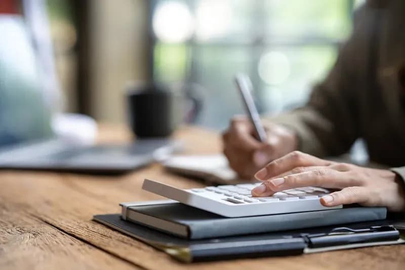 Hands using a white calculator and pen on a wooden desk over a notebook and clipboard