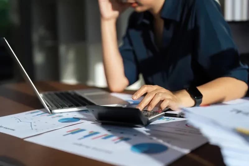Person in a dark shirt using a calculator at a desk with financial documents, charts, and a laptop, conveying concentration