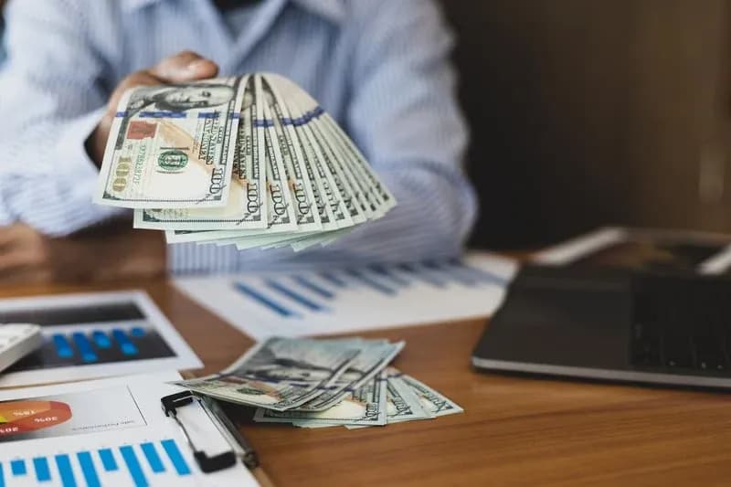 Hand holding a fan of hundred-dollar bills over a desk with financial charts and documents