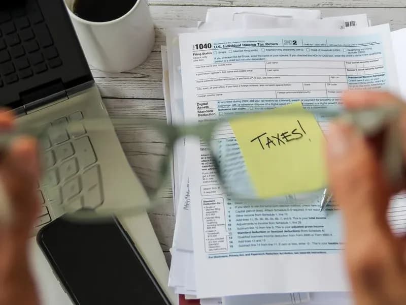 First-person view holding eyeglasses over a 1040 tax form with a yellow TAXES! sticky note, laptop and coffee on a wooden desk